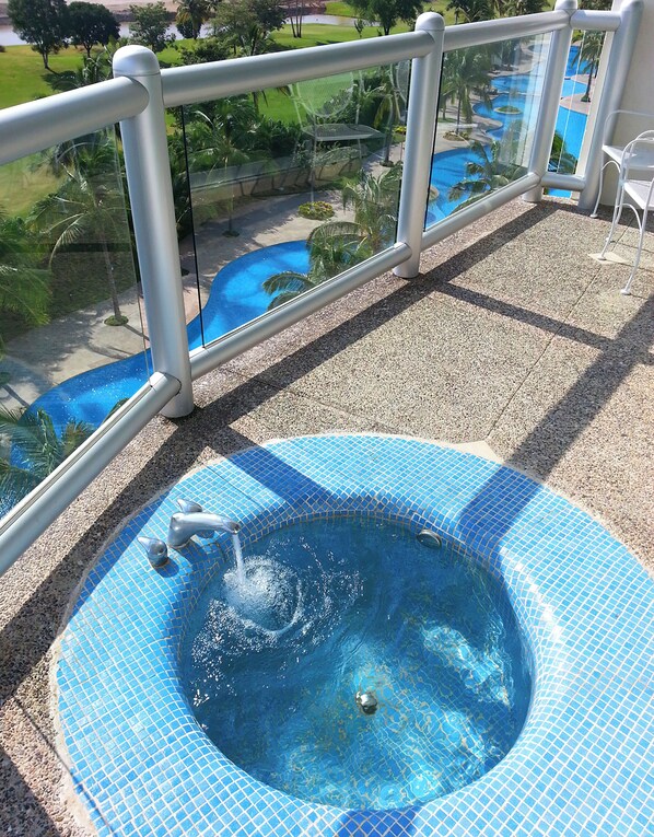 Ein Whirlpool mit einer gefliesten Kante, umgeben von einem Glasgeländer, mit Blick auf einen Pool und Grünflächen.