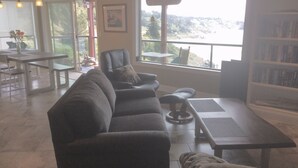 A living room with a sofa, a coffee table, and a bookshelf. There is a window with a view of a natural landscape. - A Romantic Getaway at the "White Water Cove" At Ocean View Rainbow Rock!  (Brookings)