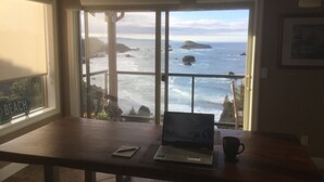 A room with a view of the ocean, a wooden desk with a laptop, a mug, and a sign indicating a beach direction. - A Romantic Getaway at the "White Water Cove" At Ocean View Rainbow Rock!  (Brookings)