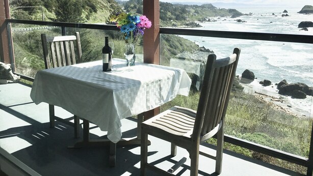 A table with a bottle and glass, a vase with flowers, and a view of the ocean and hills. - A Romantic Getaway at the "White Water Cove" At Ocean View Rainbow Rock!  (Brookings)