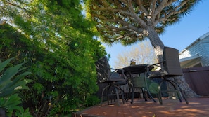 Een terras met een tafel en stoelen, omgeven door weelderige begroeiing en een grote boom.