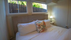 Een net opgemaakt bed met witte lakens en een decoratief kussen, een bedlampje en uitzicht op een houten omheining door een raam.