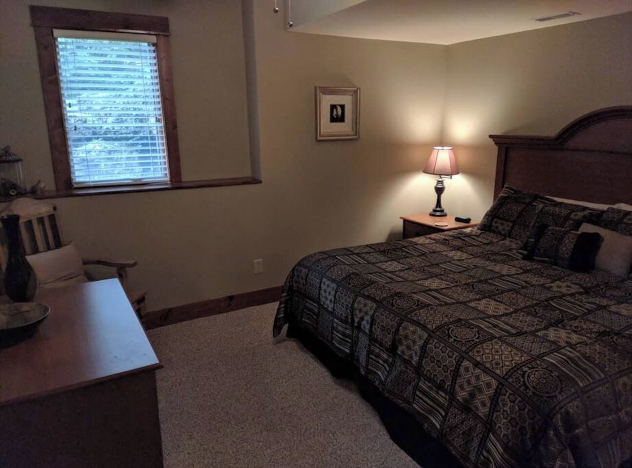 Ein Schlafzimmer mit einem Bett, einem Nachttisch, einer Lampe, einem gerahmten Bild an der Wand und einem Fenster mit Jalousien.