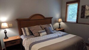 A bedroom with a bed, two bedside tables, a nightstand, a window with blinds, and a wall-mounted artwork. - Splendid Home Away From Home -5 Bdr- with Hot Tub (Galena)