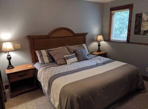 Une chambre à coucher comprenant un lit, deux tables de chevet, une commode, une fenêtre avec des stores et une œuvre d’art accrochée au mur.