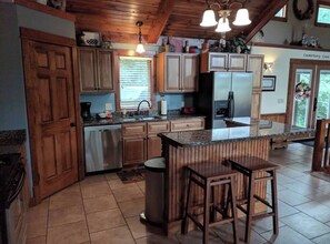 A kitchen with wooden cabinets, a stainless steel refrigerator, and a bar area with stools. - Splendid Home Away From Home -5 Bdr- with Hot Tub (Galena)