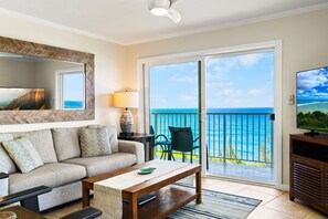 A living room with a sofa, coffee table, TV, and a balcony with a view of the ocean.