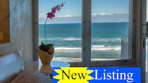 Een kamer met uitzicht op de oceaan door een groot raam, een pot orchidee op een tafel, en een bordje 'New Listing'.