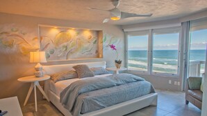 Een slaapkamer met een bed, nachtkastje, lamp, schilderij en uitzicht op de oceaan.