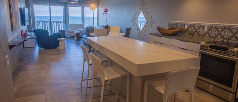 A modern kitchen with a large island, white bar stools, and a view of the ocean.