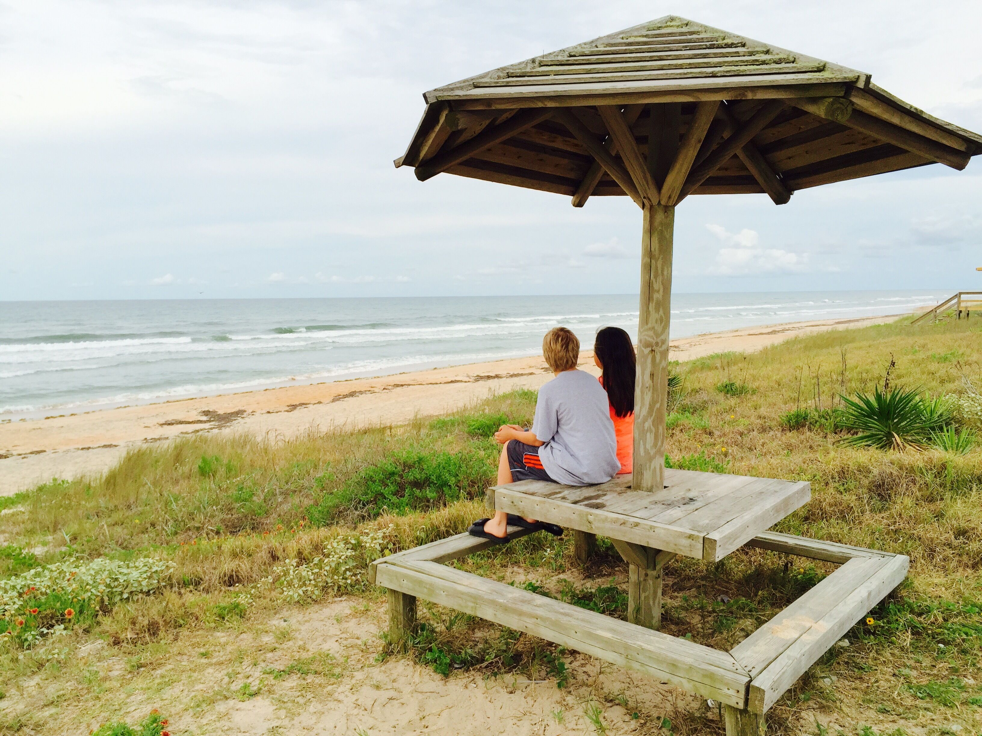 Dos personas sentadas bajo una estructura de madera en una playa, con el océano al fondo.