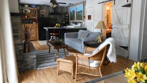 A cozy living room with a sofa, a wicker chair, a dining table, and a kitchen area with a refrigerator and a window. - WELCOME  TO THE COUNTRY - LAKE FRONT PROPERTY - THE FOX DEN STUDIO (Red Hook)