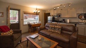 A living room with a brown leather sofa, a glass coffee table, and a dining area with a wooden table and chairs. A kitchen with white cabinets and appliances is visible in the background. - Cozy 2-Cabin Retreat Near RMNP - Estes Park License #3051 (Estes Park)