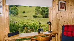 Een houten interieur met een tafel, stoelen, een lamp en een uitzicht op een groen landschap door een raam.