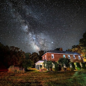 A house with a brick exterior and a covered porch, surrounded by a grassy yard and trees, under a starry night sky with visible stars and the Milky Way. - Modern 1850s Luray Farmhouse, Near Shenandoah National Park, Lake Arrowhead (Luray)