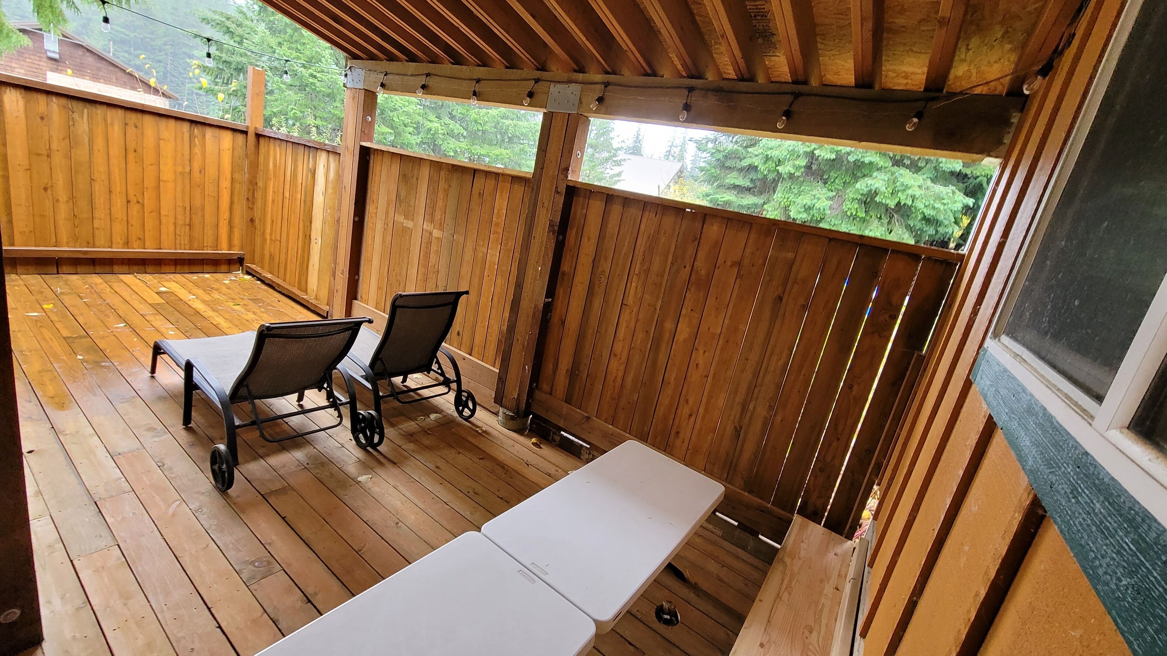 Una terraza de madera con mesa y sillas, una zona cubierta con ventanas y vistas al verdor.