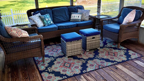 A covered porch with wicker furniture, a patterned rug, and a view of a white fence and trees. - Hayden Cottage. Great location and comfortable cottage. (Pass Christian)