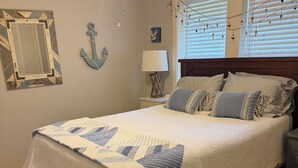 A bedroom with a bed, wooden floor, a ceiling fan, a nightstand, a lamp, and a window with blinds. - Hayden Cottage. Great location and comfortable cottage. (Pass Christian)