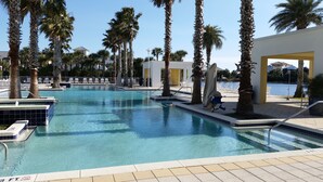 A clear blue pool surrounded by palm trees and lounge chairs, with a view of a body of water and a building in the background. - On 30A Water lovers dream!! 4 gorgeous pools, Lake and Ocean . Pet Friendly (Panama City Beach)