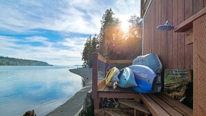 En træterrasse med kajakker og et brusehoved, med udsigt over en rolig sø med skovet bred.