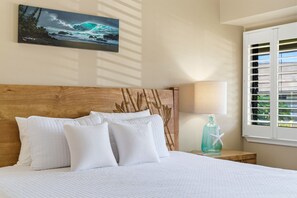 Un lit bien fait, avec une literie blanche, une tête de lit en bois ornée d’un motif de feuilles, un tableau représentant une vague accroché au mur et une fenêtre avec des stores.