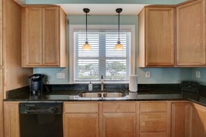 A kitchen with wooden cabinets, a black countertop, a built-in dishwasher, a double sink, and pendant lights above the window. - 🌅Oceanfront  🏖️Steps to sand 🐶Dog Friendly⚡️EV charger (North Topsail Beach)