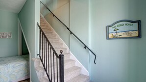 A staircase with a handrail, a bed with a patterned bedspread, and a wall sign that reads "Our Beach House." - 🌅Oceanfront  🏖️Steps to sand 🐶Dog Friendly⚡️EV charger (North Topsail Beach)