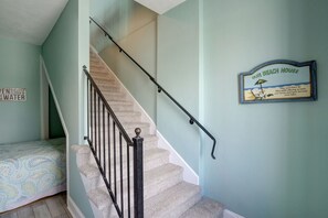A staircase with a handrail, a bed with a patterned bedspread, and a wall sign that reads "Our Beach House." - 🌅Oceanfront  🏖️Steps to sand 🐶Dog Friendly⚡️EV charger (North Topsail Beach)