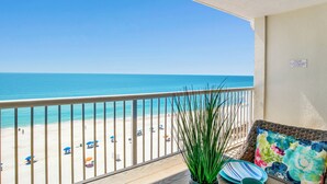 A balcony with a view of a beach, a potted plant, and a wicker chair with cushions.