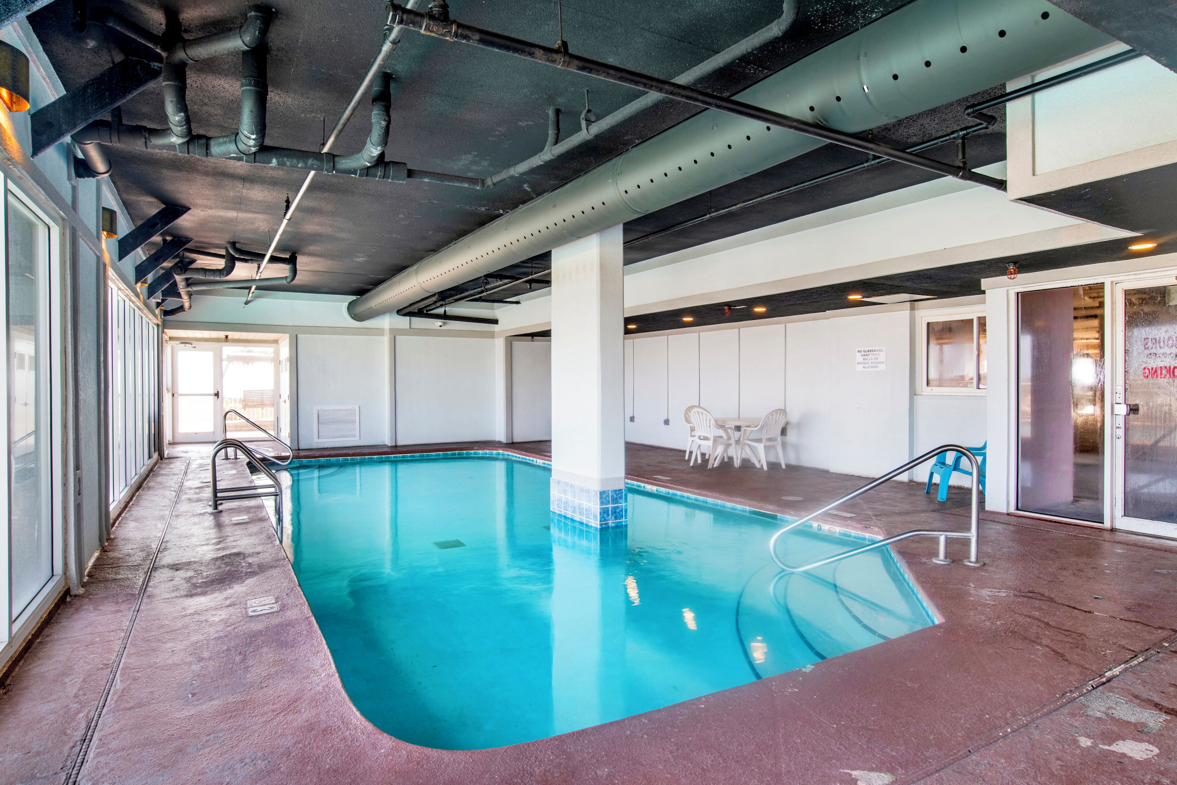 An indoor swimming pool with clear blue water, surrounded by a concrete floor and walls, with a ceiling featuring exposed pipes and ducts.