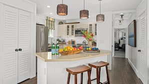 Une cuisine moderne avec des meubles blancs, un îlot central doté de tabourets, des luminaires suspendus et un panier de fruits.