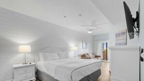 Ein Schlafzimmer mit Bett, Nachttischen, Deckenventilator, einem an der Wand befestigten Fernseher und einer Tür zu einem anderen Raum.