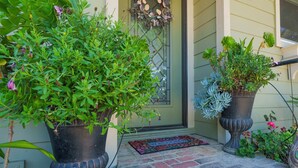 A front door with a welcome mat, two large potted plants, and a decorative wreath.