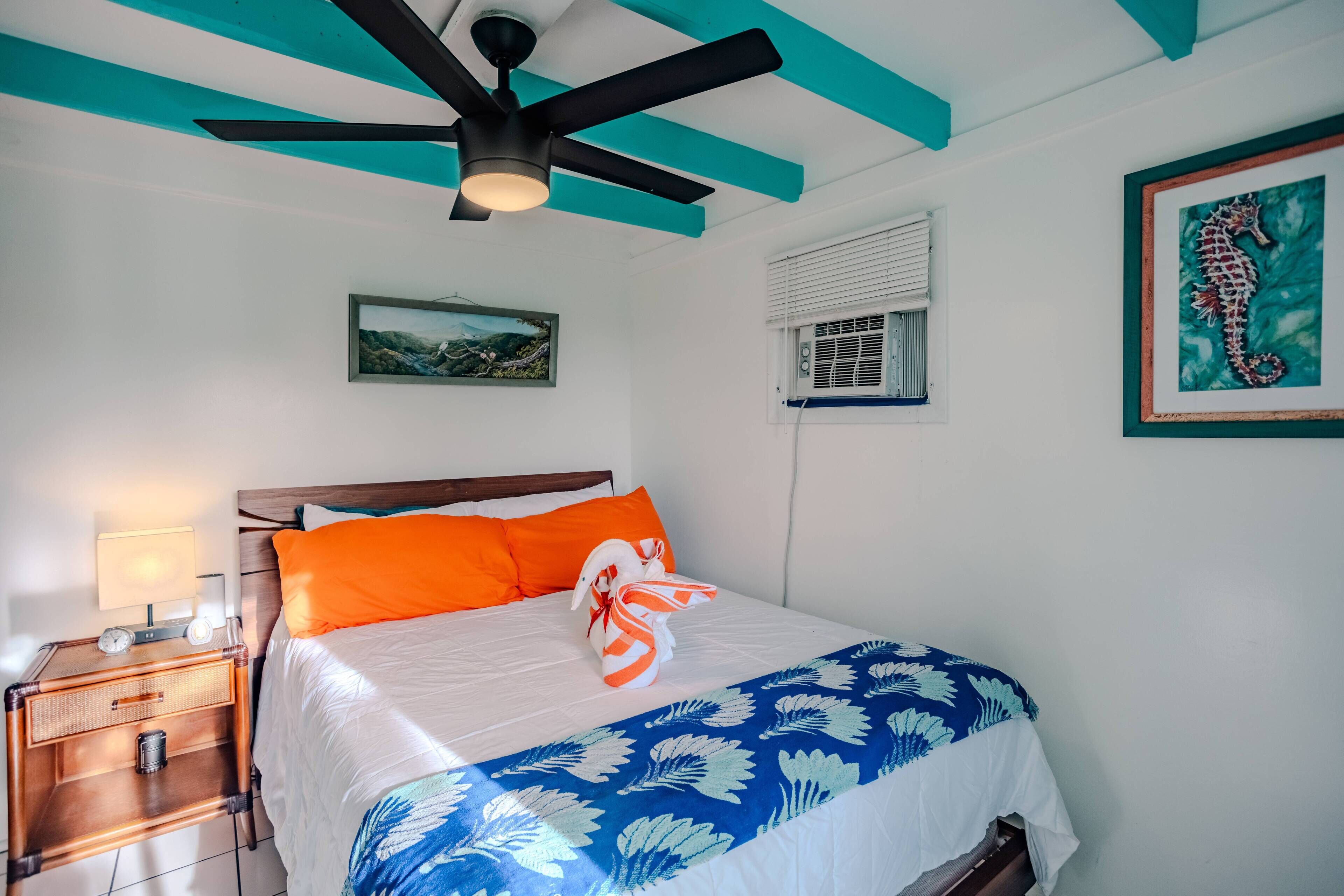 A neatly made bed with a white and blue floral bedspread, an orange pillow, and a towel shaped like a seahorse. There is a wooden nightstand with a lamp, a framed picture on the wall, and a ceiling fan.