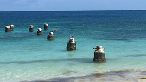 Une plage avec une eau turquoise et limpide, et plusieurs piliers en béton qui s’élèvent dans la mer.