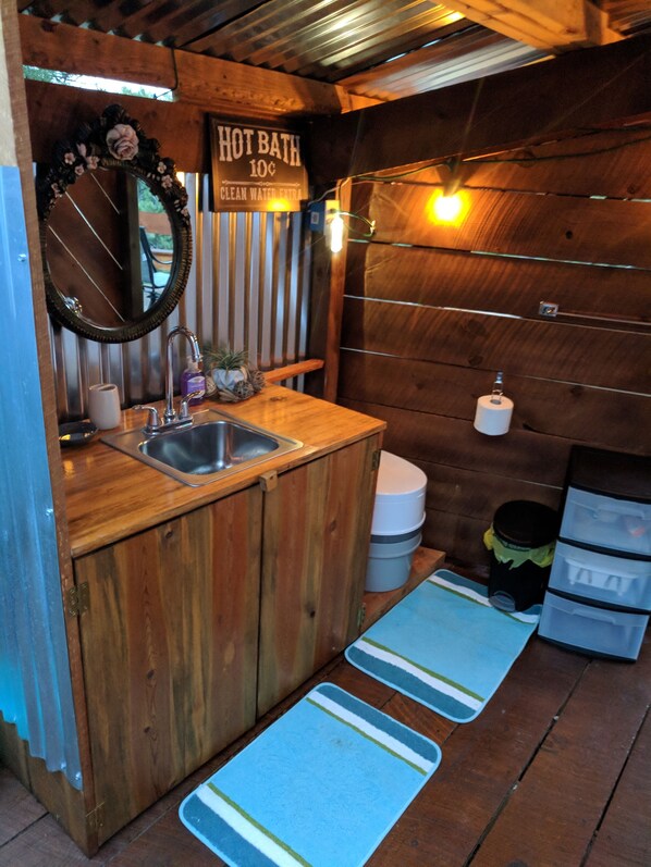 Un baño pequeño y rústico con un lavabo de madera, un espejo, un inodoro y un letrero que indica baño caliente por 10 centavos.
