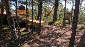 Entorno rústico al aire libre con una hamaca, terraza de madera y una silla en un área boscosa.