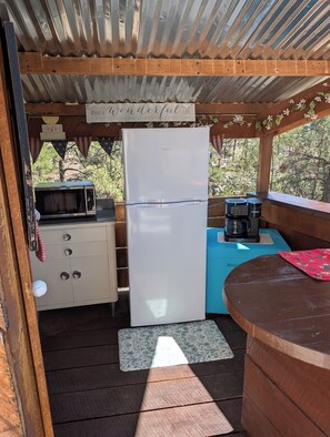 A compact kitchen with a refrigerator, microwave, and coffee maker, under a wooden structure with a view of trees.