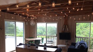 L’intérieur d’une cabane est doté d’un plafond en bois, de guirlandes lumineuses, d’un coin repas avec une table et des chaises, d’un téléviseur fixé au mur et offre une vue sur l’extérieur grâce à de grandes fenêtres.