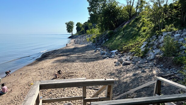 Une terrasse en bois mène à une plage de sable, avec une colline rocheuse et des arbres en arrière-plan.
