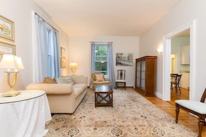 A living room with a sofa, armchairs, a coffee table, and a patterned rug. There is a dining area visible through an open doorway.