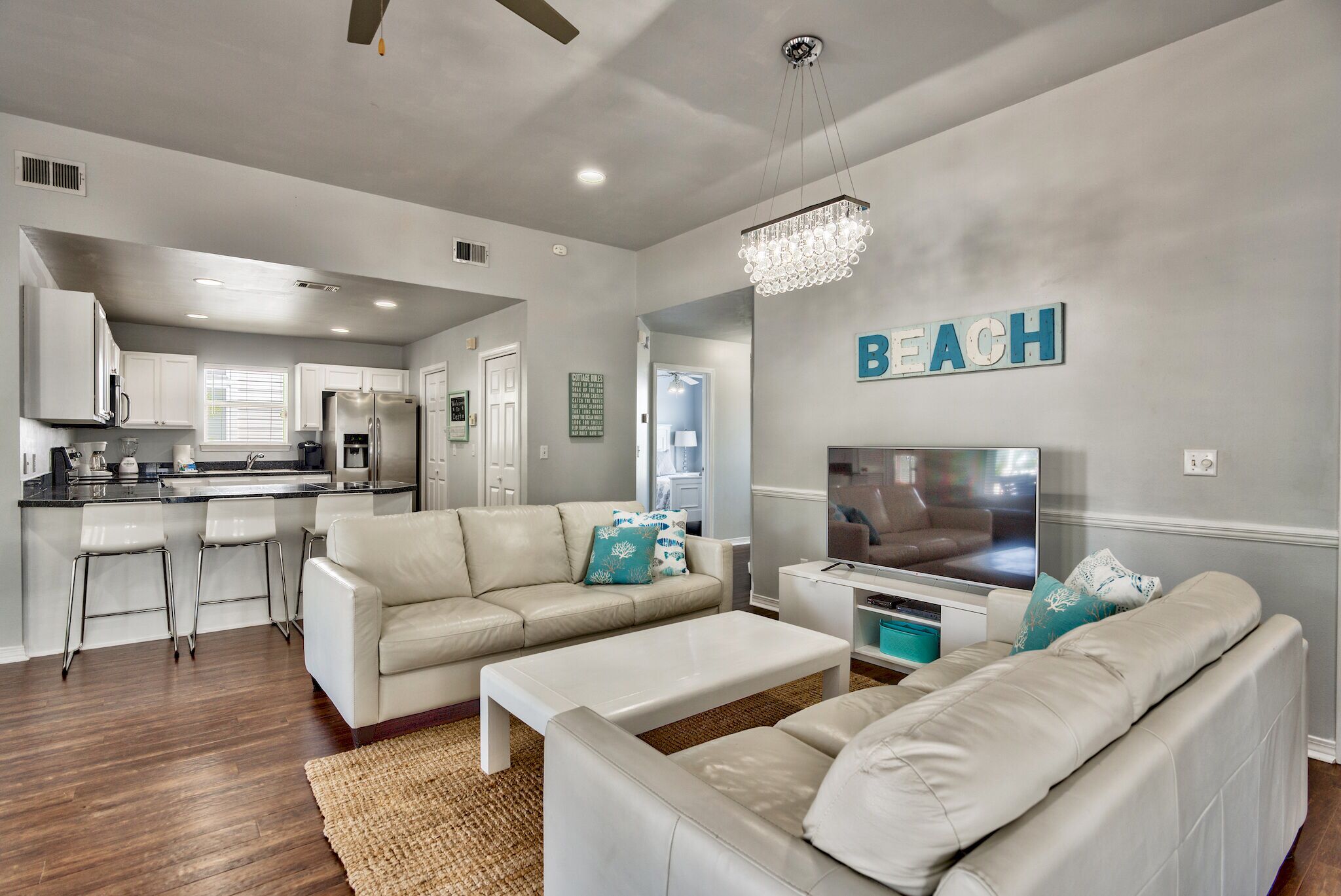 A modern living room with a white sofa set, a coffee table, and a TV. There is a kitchen area in the background with white cabinets and a stainless steel refrigerator.