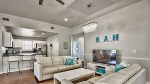 A modern living room with a white sofa set, a coffee table, and a TV. There is a kitchen area in the background with white cabinets and a stainless steel refrigerator.