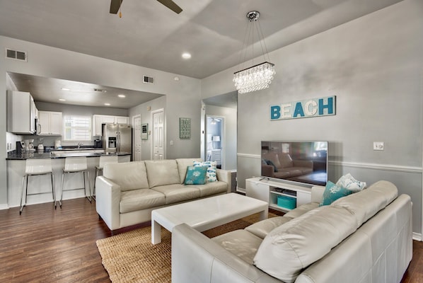 A modern living room with a white sofa set, a coffee table, and a TV. There is a kitchen area in the background with white cabinets and a stainless steel refrigerator.