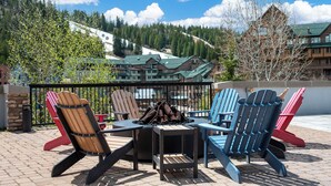 A patio with Adirondack chairs and a fire pit, overlooking a mountain lodge.