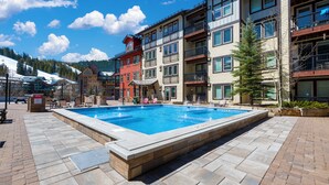 A swimming pool with a stone border, surrounded by multi-story buildings with balconies and a view of a snowy mountain.