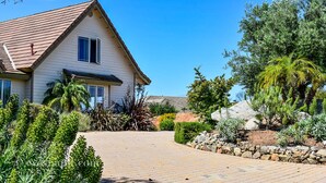 A single-story house with a tiled roof, surrounded by a variety of plants and a stone pathway leading to the entrance. - San Diego Country Barndominium, Mountain Views, Spa, Deck, & More. (Valley Center)