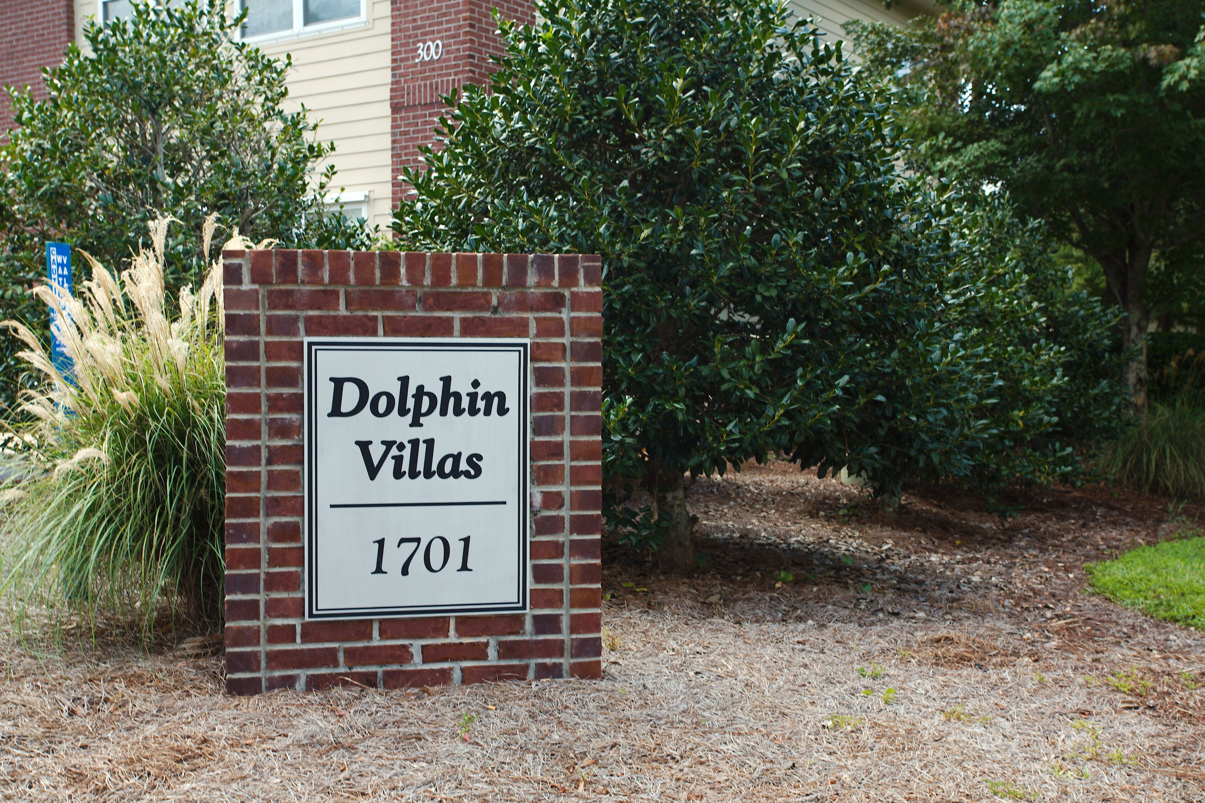 A sign for Dolphin Villas with the address 1701, surrounded by greenery and a brick building in the background.