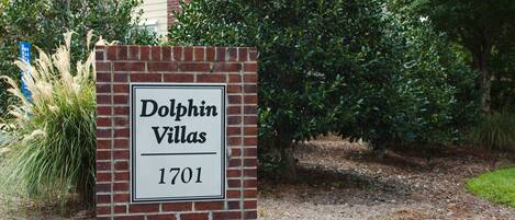 A sign for Dolphin Villas with the address 1701, surrounded by greenery and a brick building in the background.