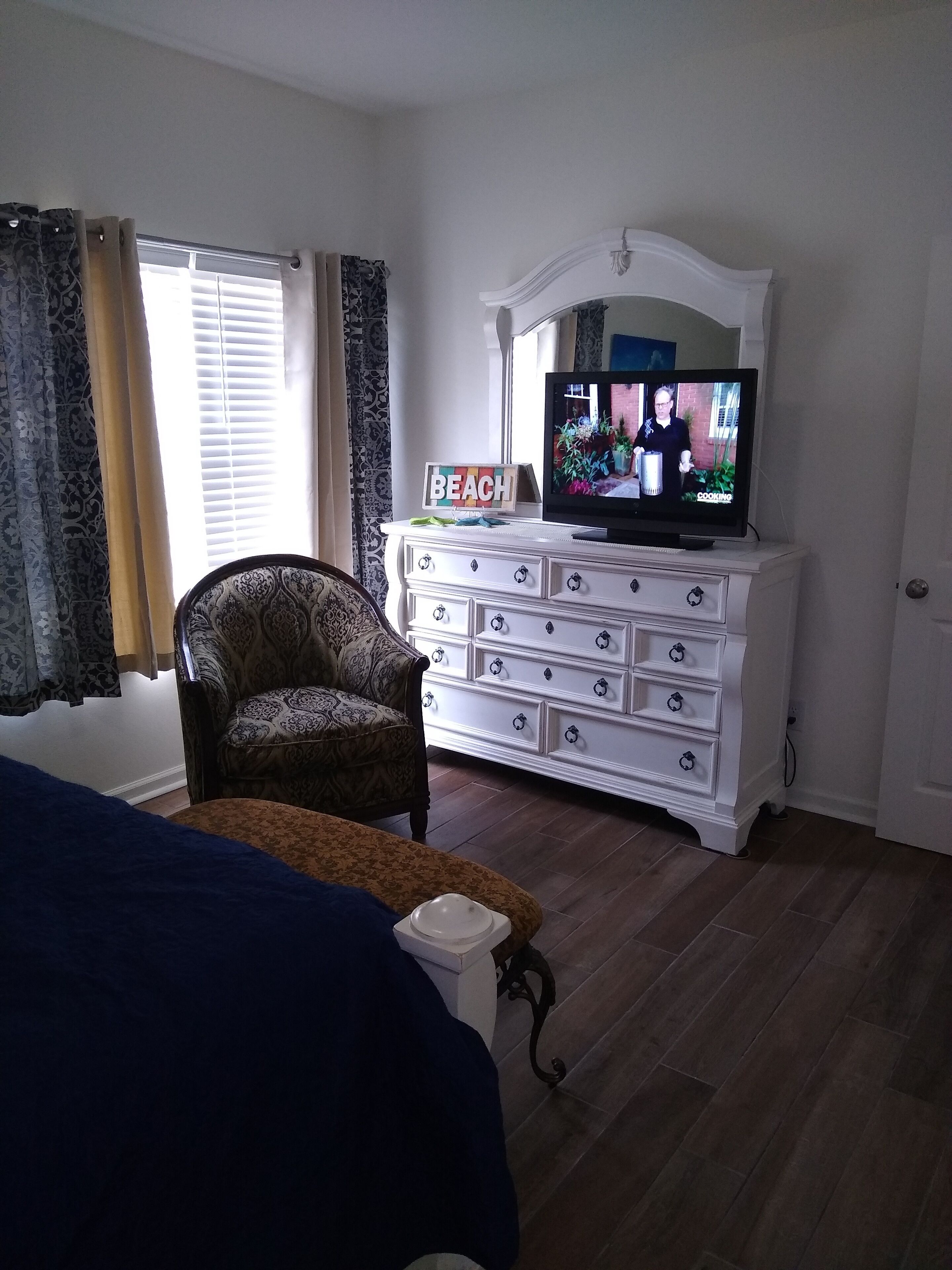 Una habitación con una cama, un televisor sobre una cómoda, una silla y un ventanal con cortinas.